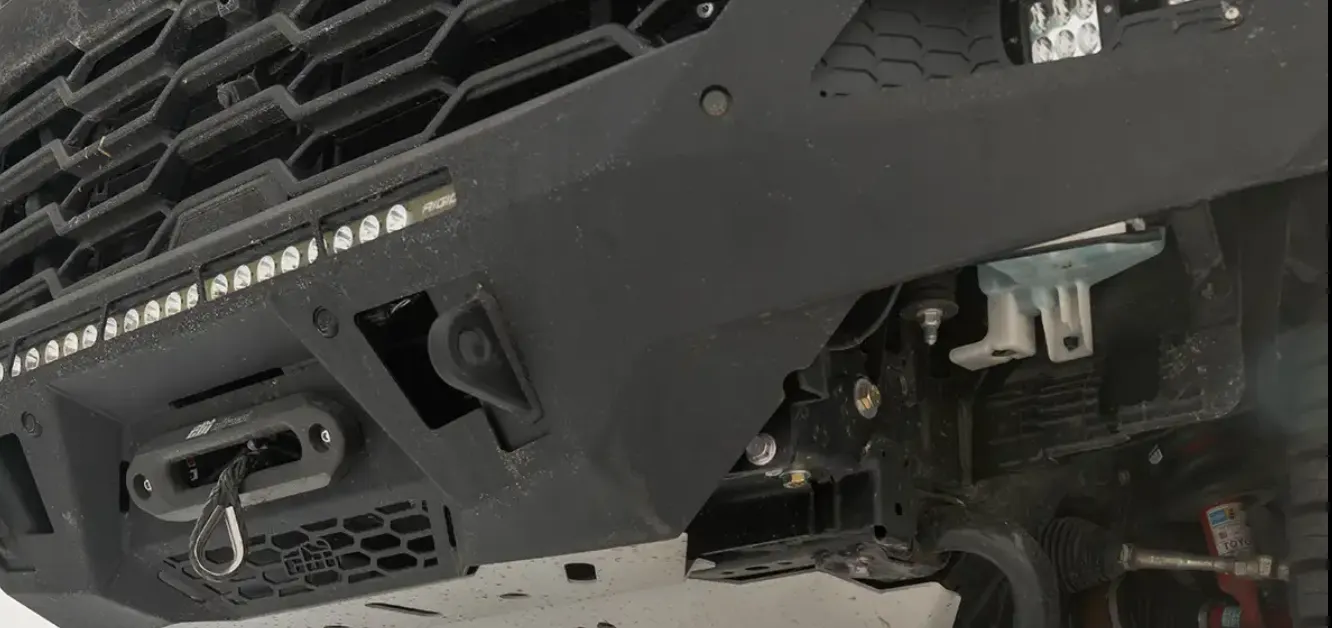 Close-up of a matte black steel off-road front bumper with an integrated LED light bar, winch fairlead, and aluminum skid plate
