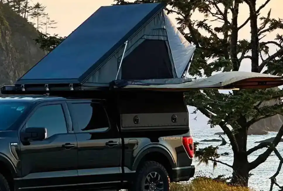 Black pickup truck with a hard camper shell, rooftop tent, and deployed awning parked on a coastal bluff