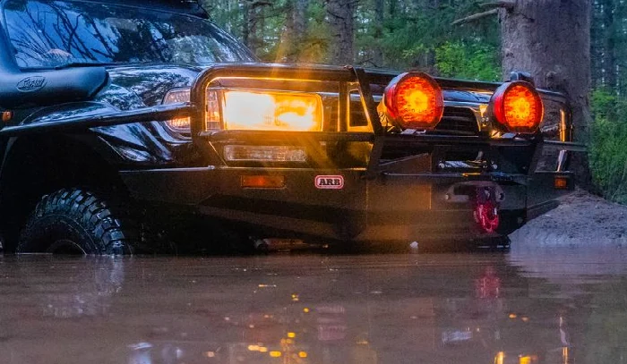 Black off-road truck with an ARB bull bar, Safari snorkel, and auxiliary lights crossing deep water on a forest trail